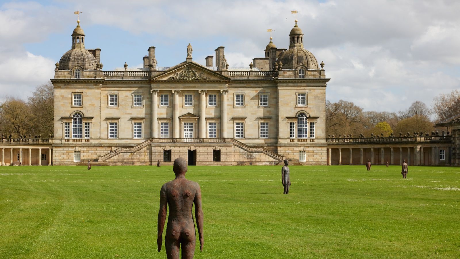 A large stately home and its grounds are pictured on a sunny day. A series of bronze figures are arranged on the grass.