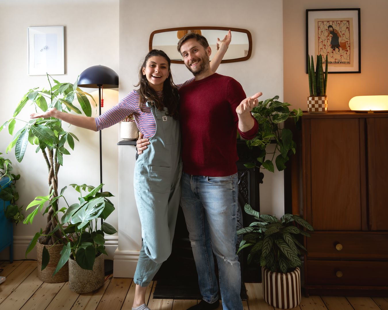 A woman wearing dungarees and a man wearing a red jumper stand with their arms outstretched in front of a fireplace and a group of houseplants