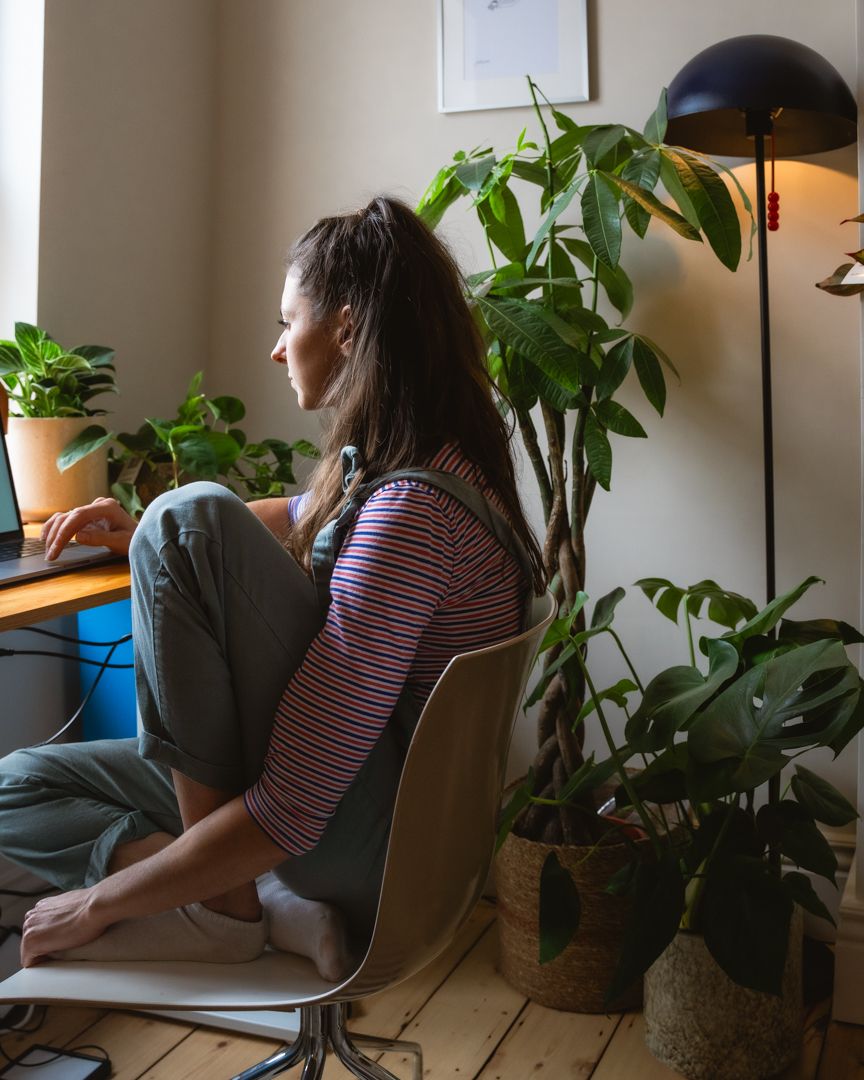 A woman wearing dungarees sits at a desk surrounded by plants