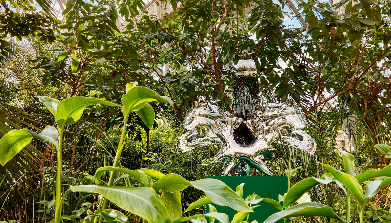 A silver reflective sculpture of an orchid is surrounded by plants in a large greenhouse.