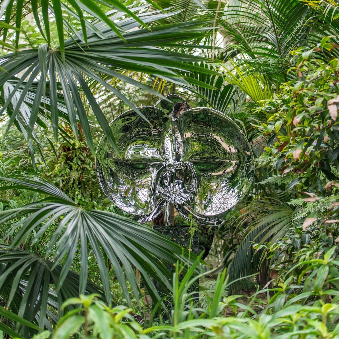 A silver reflective sculpture of an orchid is surrounded by plants in a large greenhouse.